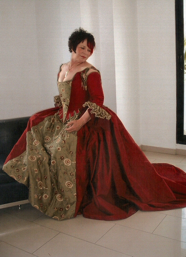 The main corset and skirt were constructed in a deep red/ black silk dupion with the contrasting embroidered fabric used as a stomacher front to the bodice and an apron front to the skirt. 
A hooded cloak, shown in a soft sage green velvet, was designed to tone with the 18th century style gown. It was lined in red silk dupion.  The double sleeve falls of lace and organza at the cuff give an authentic 18th century feel.