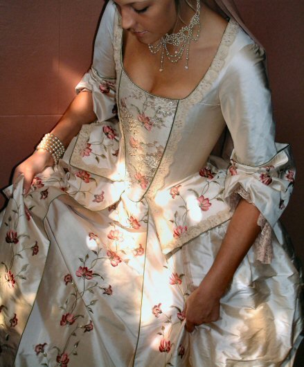 The bodice is ornamented with an eye-catching embroidered and beaded stomacher panel and finishes in a shaped and pleated peplum, embroidered and edged in antique lace. The bodice is laced at the back to achieve a corset-like finish. It has fitted, elbow-length sleeves ending in large, romantic cuffs with lace sleeve flounces. The skirt has an open front to show off the beautiful scrolling floral embroidery and draped panniers, again edged in lace.  Shown in a pale pink embroidered regal dupion, identical fabric is also available in a buttermilk yellow, french grey/ blue, ivory and taupe/cream.  All with toning embroideries. - £3000  This gown can also be constructed in any other dupion, taffeta, zibilene or thai silk with appropriate trims - £2200+. Please enquire for quotes on all variations.